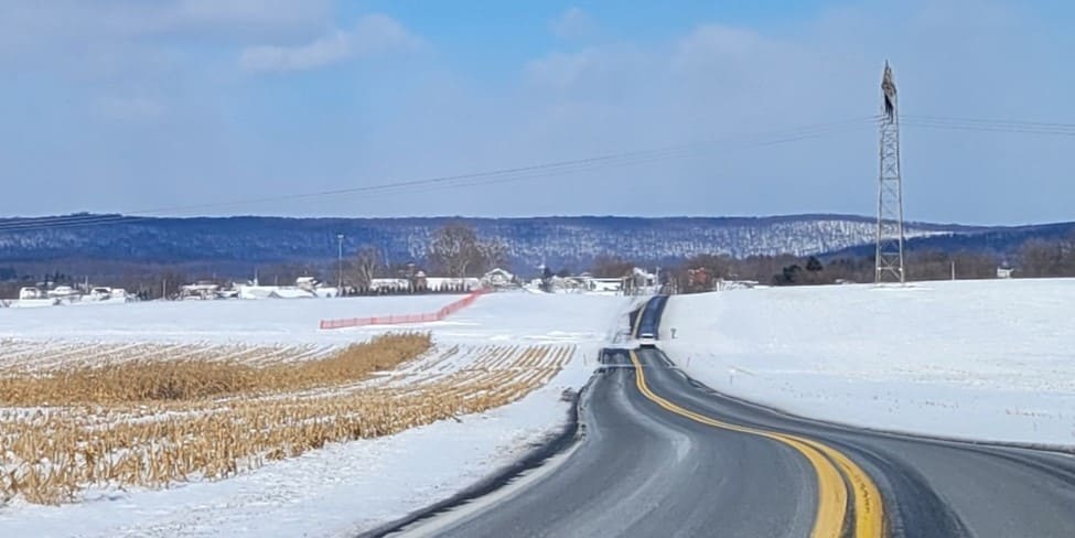 An image of a rural, two-lane roadway, with snow-covered cornstalks on the left-hand side and an open field on the right-hand side.